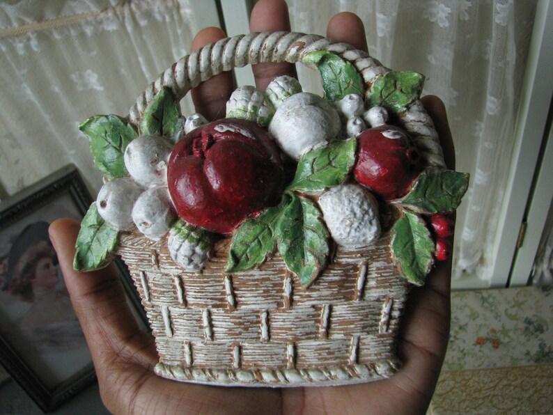 May include: A white ceramic basket filled with red and white fruit, with green leaves. The basket is decorated with a rope-like trim.
