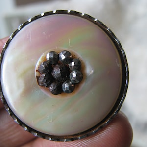 Puede incluir: Un botón redondo de nácar blanco con un borde plateado y un grupo de piedras negras de vidrio en el centro.