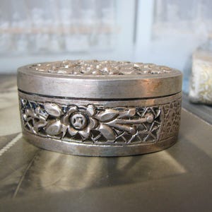 May include: A silver-toned, oval-shaped trinket box with intricate floral and geometric patterns. The lid features a textured surface, while the sides display detailed openwork designs. This decorative box is likely used for storing small items.