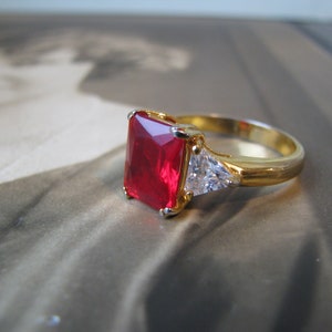 May include: A gold ring with a large, square-shaped red gemstone and two smaller, triangular clear gemstones.
