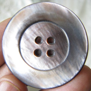 May include: A large, round, four-hole, mother-of-pearl button with a dark gray iridescent sheen.