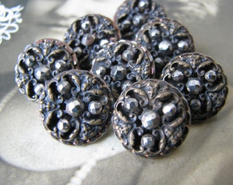 May include: A collection of antique-style buttons. Each button is round, with an ornate, bronze-colored metal design featuring swirling patterns and faceted, silver-colored accents. The buttons are clustered together, showcasing their intricate details.