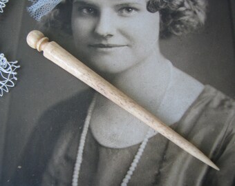 May include: A light-colored wooden hair pin with a rounded top and a tapered, pointed end. The pin is set against a vintage black and white photograph of a woman. The hair accessory is a classic design.