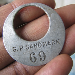 May include: A silver metal tag with the text "S.P. SANDMARK 69" engraved on it.