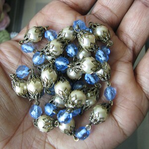 May include: A white and blue beaded necklace with a decorative metal design. The necklace is made of round white beads with a metal frame and blue glass beads.