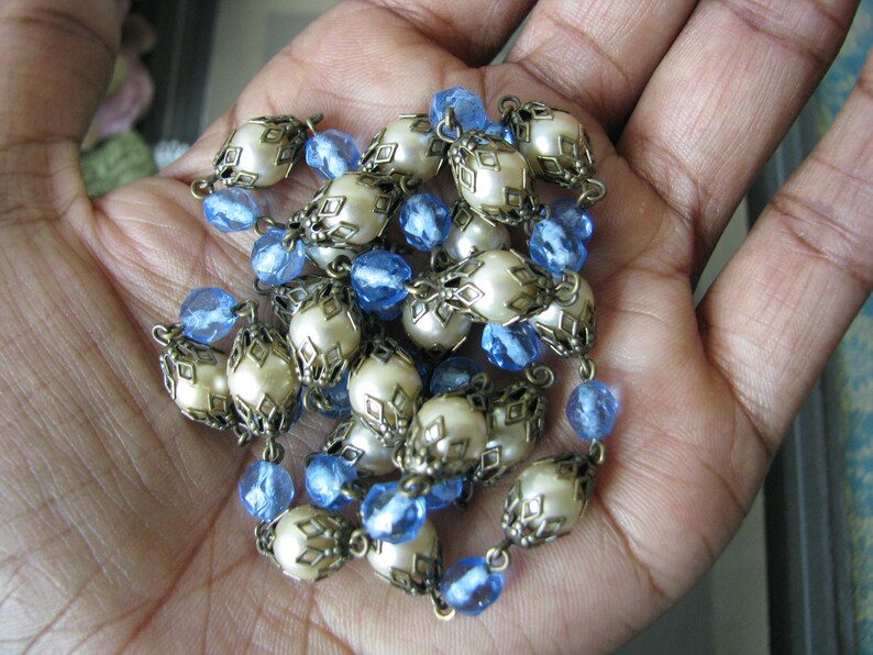May include: A white and blue beaded necklace with a decorative metal design. The necklace is made of round white beads with a metal frame and blue glass beads.