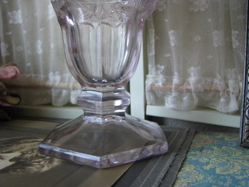 Puede incluir: Un jarr&oacute;n de vidrio transparente con un delicado patr&oacute;n floral y una base hexagonal. El jarr&oacute;n tiene un tono lavanda p&aacute;lido.