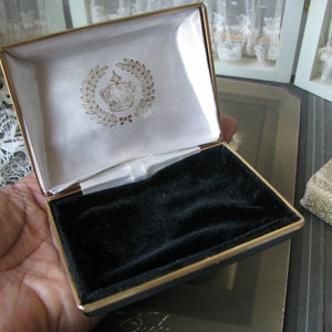 May include: A gold-trimmed, black velvet-lined jewelry box with a white satin interior. The lid features a gold crown with a laurel wreath design.