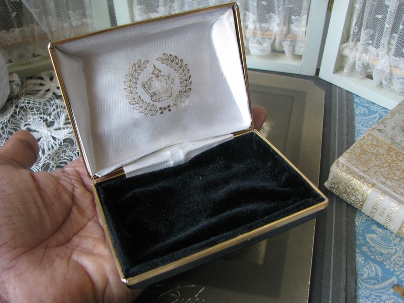 May include: A gold-trimmed, black velvet-lined jewelry box with a white satin interior. The lid features a gold crown with a laurel wreath design.
