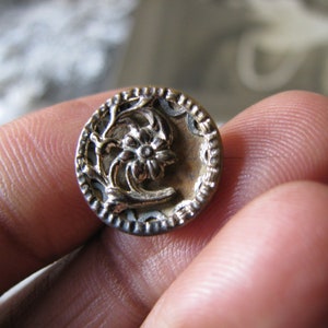 Puede incluir: Un botón plateado con un diseño floral. El botón tiene un borde elevado y adornado.