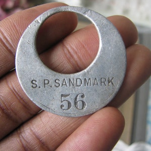 May include: A silver metal tag with the text "S.P. SANDMARK 56" engraved on it.
