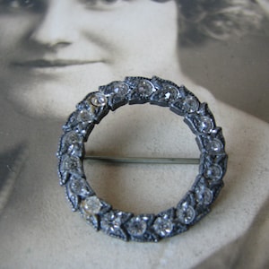 Puede incluir: Un broche plateado en forma de círculo, decorado con diamantes de imitación transparentes engastados en un diseño de hojas.