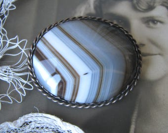 May include: Oval brooch with a polished agate stone featuring bands of white, brown, and blue-gray. The stone is set in a silver-toned, twisted rope-like frame. The background includes a vintage photograph and white lace.