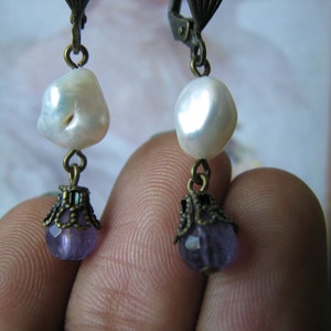 Puede incluir: Un par de pendientes con una perla blanca y una amatista púrpura. Los pendientes están hechos de metal bronce y tienen un diseño delicado.