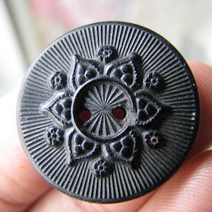 Puede incluir: Un botón de plástico negro con un diseño floral decorativo y un patrón radial alrededor de los agujeros centrales.