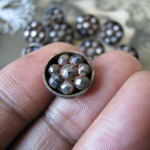 Op de afbeelding: Een close-up van een zilveren metalen knoop met een cirkelvormig ontwerp. De knoop heeft een verhoogd midden met acht kleine, ronde, zilveren studs. De knoop is omgeven door een zilveren metalen rand.