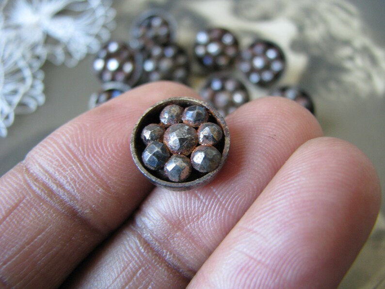 Op de afbeelding: Een close-up van een zilveren metalen knoop met een cirkelvormig ontwerp. De knoop heeft een verhoogd midden met acht kleine, ronde, zilveren studs. De knoop is omgeven door een zilveren metalen rand.