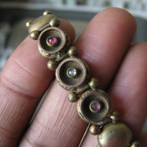 Puede incluir: Un brazalete dorado con eslabones redondos decorativos. Cada eslabón presenta un círculo elevado con una pequeña piedra transparente o rosa en el centro.