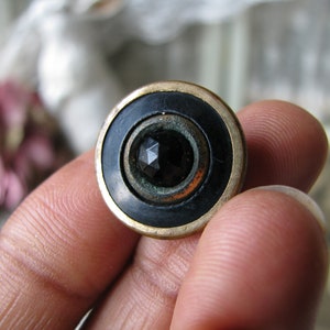 May include: A black and gold button with a faceted black glass center. The button has a raised rim and a smooth, shiny finish.