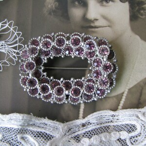 May include: A silver oval brooch with a decorative design of purple rhinestones. The brooch is set against a white lace background.