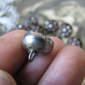 Op de afbeelding: Een zilveren metalen knoop met een ronde, gewelfde bovenkant en een kleine lus aan de achterkant.