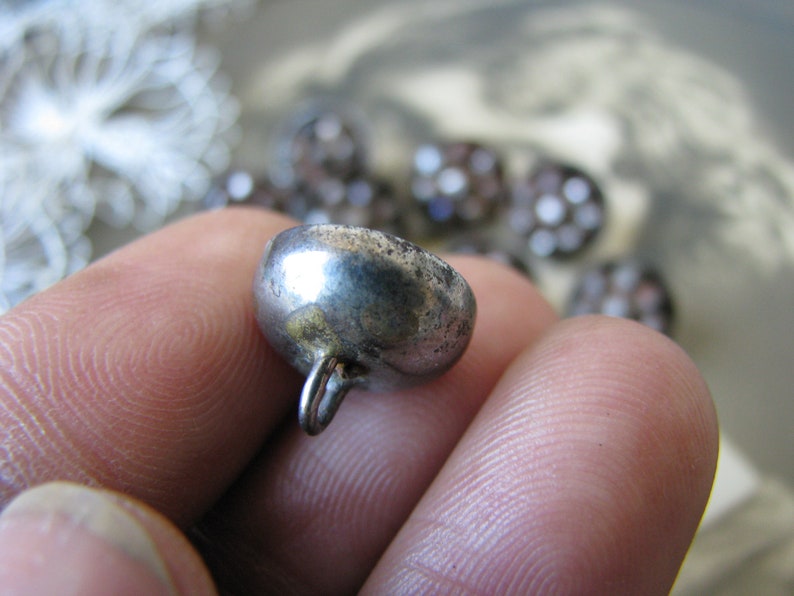 Op de afbeelding: Een zilveren metalen knoop met een ronde, gewelfde bovenkant en een kleine lus aan de achterkant.