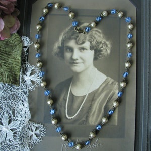 May include: A vintage photo of a woman wearing a pearl necklace and a silver and blue beaded necklace. The photo is framed and displayed on a table with a lace doily and a purple flower.