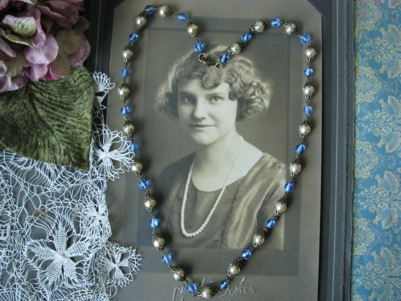 May include: A vintage photo of a woman wearing a pearl necklace and a silver and blue beaded necklace. The photo is framed and displayed on a table with a lace doily and a purple flower.