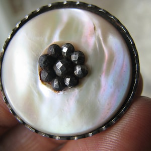 Puede incluir: Un botón de nácar blanco con un borde plateado y un grupo de tachuelas de metal negro en el centro.