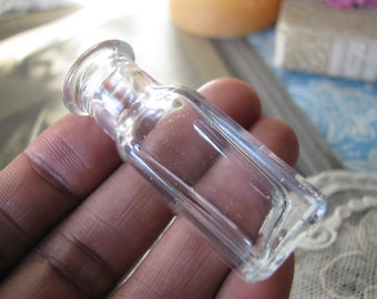 Puede incluir: Una pequeña botella de vidrio transparente con cuerpo rectangular y cuello redondeado. La botella se sostiene en una mano, mostrando su tamaño y forma. El vidrio parece ser vintage o antiguo.