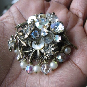Puede incluir: Un broche vintage con un grupo de piedras de cristal transparentes e iridiscentes, perlas falsas y flores de metal. El broche es de color dorado y tiene una cadena colgante de perlas y piedras de cristal.