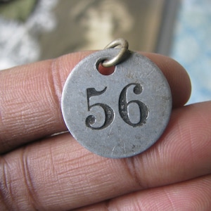 May include: A silver metal tag with the number 56 engraved on it. The tag has a hole at the top for attaching it to something.