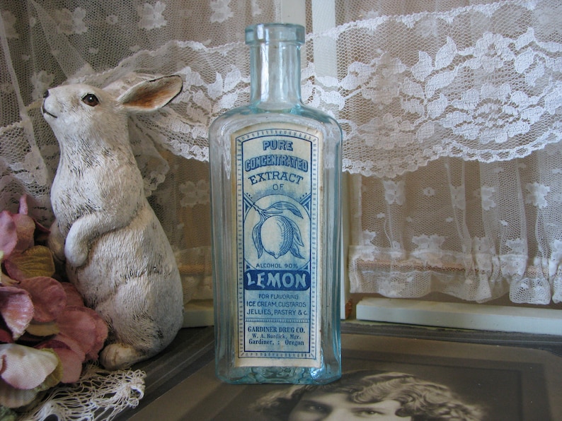 May include: A vintage glass bottle with a blue and white label that reads "Pure Concentrated Extract of Lemon, Alcohol 90%, For Flavoring Ice Cream Custards Jellies, Pastry &c, Gardiner Drug Co., W.A. Barrick, Mgr., Gardiner, Oregon".