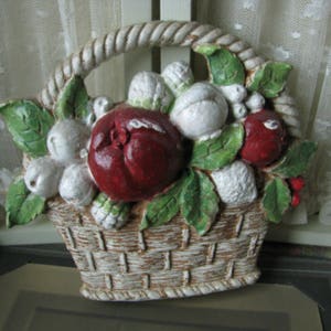 May include: A white ceramic basket filled with red and white fruit. The basket has a rope handle and is decorated with green leaves.