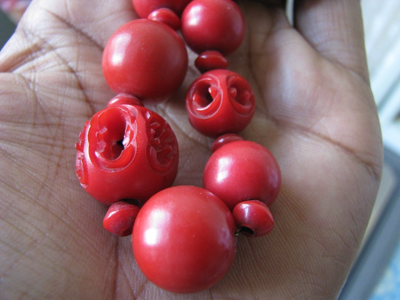 Peut inclure: Un collier de perles rouges avec des d&eacute;tails sculpt&eacute;s. Les perles sont rondes et lisses, et le collier est enfil&eacute; sur un cordon.
