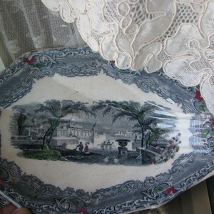 May include: A blue and white oval serving dish with a decorative border. The center of the dish features a black and white print of a landscape with trees and buildings.