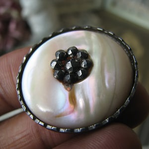 Puede incluir: Un botón de nácar blanco con un borde plateado y un grupo de diamantes de imitación negros en el centro.