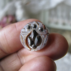 Puede incluir: Un botón blanco y marrón con un diseño floral y detalles plateados. El botón tiene un diseño en relieve y está hecho de un material liso.