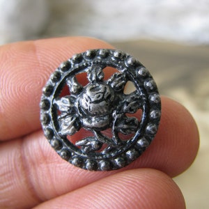Puede incluir: Un botón de metal plateado con un diseño en relieve detallado de una rosa con hojas. El botón tiene forma circular con un borde de cuentas.