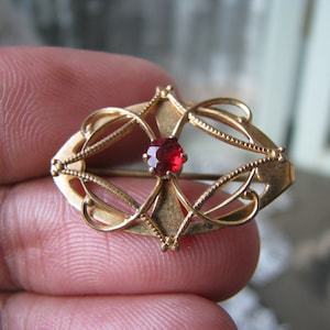 Pode incluir: Um broche dourado com uma pedra vermelha no centro. O broche é desenhado com linhas intrincadas e em espiral.