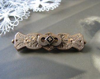 May include: A gold-toned brooch with an intricate floral design and a black enamel center featuring a cross. The brooch is in the shape of an infinity symbol.
