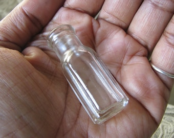 Puede incluir: Una botella de vidrio transparente con forma cuadrada y superficie texturizada. La botella está vacía y tiene una pequeña abertura en la parte superior.