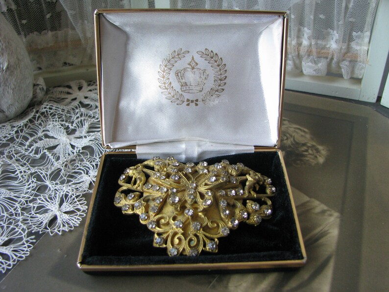 May include: A gold-tone brooch in the shape of a butterfly with clear rhinestones. The brooch is in a black velvet-lined box with a white satin interior. The box has a gold crown logo with a laurel wreath.
