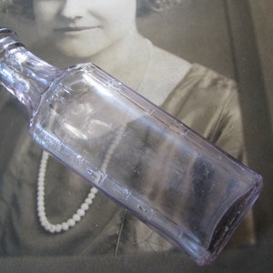May include: A clear glass bottle with a light purple tint. The bottle has a rounded bottom and a long, narrow neck. The bottle is empty and has a textured surface.