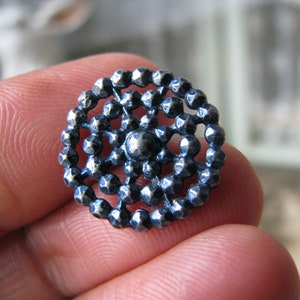 May include: A silver-toned button with a circular design featuring a central raised dot surrounded by concentric circles of smaller raised dots.
