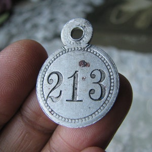 May include: A vintage silver metal tag with the number "213" embossed on its face. The tag has a circular shape with a beaded border and a small loop at the top for hanging. The metal has a worn, aged appearance.