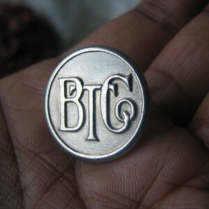May include: A silver-colored metal button with the letters "BTC" embossed on it.