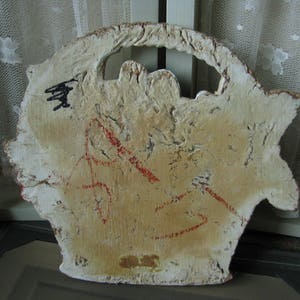 May include: A white wooden plaque with a cutout design and red markings. The plaque has a rough, textured surface and appears to be vintage.