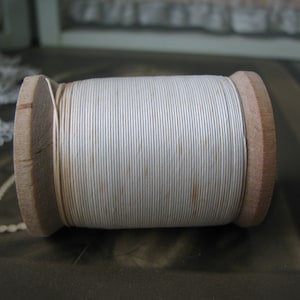 Op de afbeelding: Een spoel wit garen gewikkeld om een houten spoel. Het garen is glad en heeft een lichte glans.