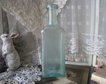 Puede incluir: Una botella de vidrio azul claro, rectangular y con cuello estrecho. La botella tiene un aspecto ligeramente descolorido, lo que sugiere que es antigua. El fondo incluye una cortina de encaje blanco y un conejo de cerámica.